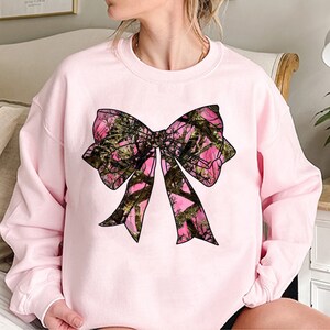 a woman sitting on a bed wearing a pink sweatshirt with a bow on it