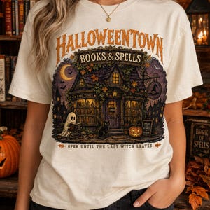 May include: A cream-coloured t-shirt featuring a graphic of a bookstore titled HALLOWEENTOWN BOOKS & SPELLS. The illustration depicts a shop with a ghost, a black cat, a pumpkin, and the text OPEN UNTIL THE LAST WITCH LEAVES.