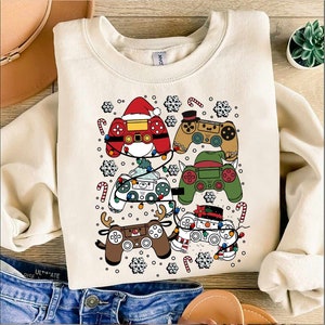 May include: A white sweatshirt with a Christmas-themed design featuring multiple video game controllers decorated with festive elements like candy canes, snowflakes, and Christmas lights.