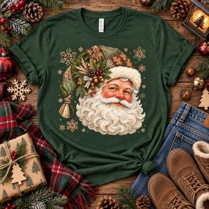 May include: A dark green t-shirt featuring a graphic of a smiling Santa Claus with a long white beard. Santa wears a patchwork-style hat decorated with pine needles, acorns, and berries. The design is surrounded by small, light-coloured snowflake icons.
