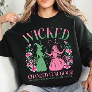 Könnte beinhalten: Schwarzer Pullover mit einem pinkfarbenen und grünen Motiv von zwei singenden Frauen. Der Text "Wicked Changed for Good" ist unter dem Motiv gedruckt. Der Text "Because I knew you have been changed for good" ist unter dem Haupttext gedruckt.