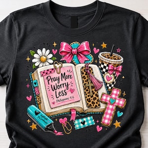 May include: A black t-shirt featuring a graphic of an open book with the text "Pray More Worry Less Philippians 4:6". The design includes a pink bow, a leopard print bookmark with a cross, a checkered cup, a daisy, and a blue highlighter.