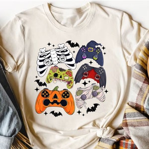 Op de afbeelding: Een wit T-shirt met een ontwerp van verschillende Halloween-videogamecontrollers. De controllers zijn versierd met skeletten, zombies, mummies en pompoenen.