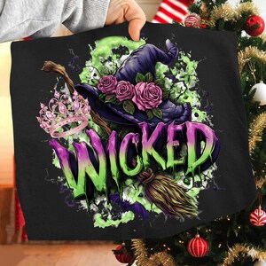 May include: Black graphic featuring a witch's hat with pink roses, a pink crown, and a broom. The word "WICKED" is displayed in a green and purple gradient. A green and white splatter effect surrounds the design.