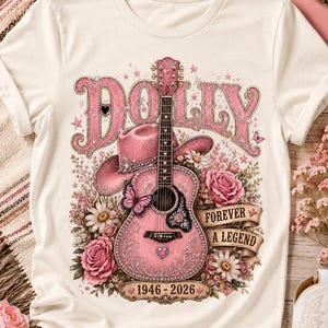 Può includere: Una t-shirt color crema con una stampa grafica di una chitarra acustica rosa, un cappello da cowboy rosa, rose rosa e margherite. Il testo recita DOLLY, FOREVER A LEGEND e 1946-2026. Il design include una farfalla rosa e dettagli a stella.