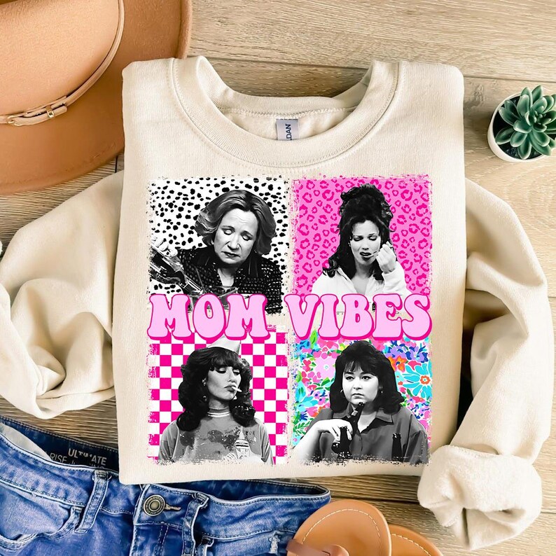 a white sweatshirt with a graphic design that includes four black and white photographs of women, along with the text "MOM VIBES" in pink and white.