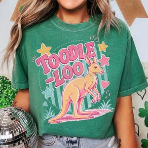 Può includere: T-shirt verde con grafica di canguro e la scritta "Toodle Loo" in rosa. Il canguro è arancione chiaro con dettagli rosa, circondato da stelle e cuori. Una t-shirt casual e unisex.