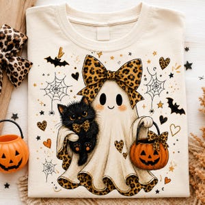 May include: A cream-colored t-shirt featuring a graphic of a ghost wearing a large leopard print bow, holding a black kitten with a matching bow, and carrying a jack-o'-lantern bucket. The design includes spiderwebs, bats, and hearts.