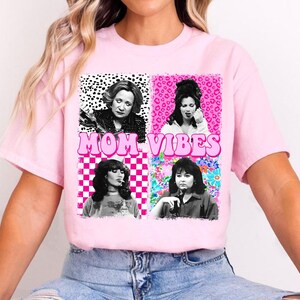 The image is a pink t-shirt with a graphic design featuring four black and white photographs of women, along with the text "MOM VIBES" in pink letters.