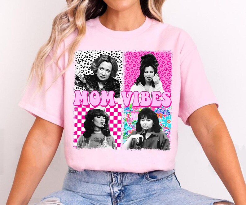 The image is a pink t-shirt with a graphic design featuring four black and white photographs of women, along with the text "MOM VIBES" in pink letters.