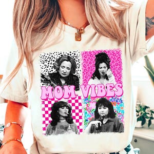 a woman wearing a white t-shirt with a graphic design that includes four different portraits of a woman, along with the text "MOM VIBES" prominently displayed.