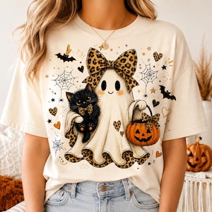 A woman wearing a white t-shirt with a Halloween-themed graphic design featuring a ghost, a cat, a pumpkin, and various Halloween symbols.