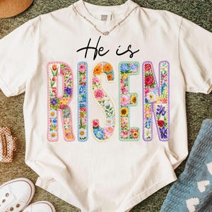 Op de afbeelding: Crèmekleurig T-shirt met de woorden "He is Risen" in zwart schrift. De letters van "Risen" zijn gevuld met kleurrijke bloemenpatronen, waaronder tulpen, rozen en zonnebloemen. Een ketting is zichtbaar bij de halslijn.