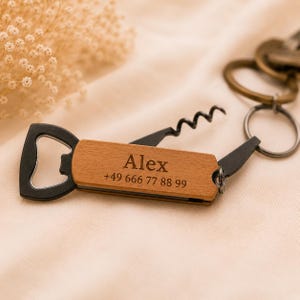 Könnte beinhalten: Ein Multi-Tool-Schlüsselanhänger mit einem Holzgriff, in den der Name Alex und die Telefonnummer +49 666 77 88 99 eingraviert sind. Das Werkzeug umfasst einen schwarzen Flaschenöffner aus Metall und einen Korkenzieher an einem Metallring.