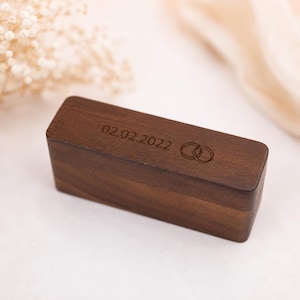 May include: A rectangular dark brown wooden box with a smooth finish. The top surface features a laser-engraved design consisting of the date 02.02.2022 followed by an icon of two interlocking rings, positioned horizontally on the lid.