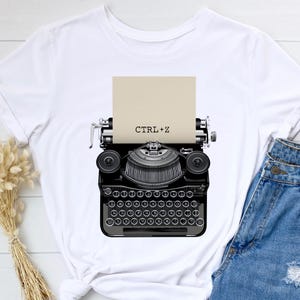 May include: White t-shirt with a black and white illustration of a vintage typewriter with a piece of paper that says "CTRL+Z".