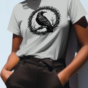 May include: A light gray t-shirt featuring a black and white illustration of a raven perched on a branch within a circular thorny wreath. The raven is detailed with visible feathers and a curved beak. The shirt is made of a soft material.
