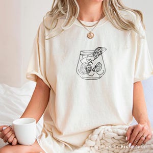 May include: A white t-shirt with a black line drawing of a glass of lemonade with lemon slices. The shirt is being worn by a person.