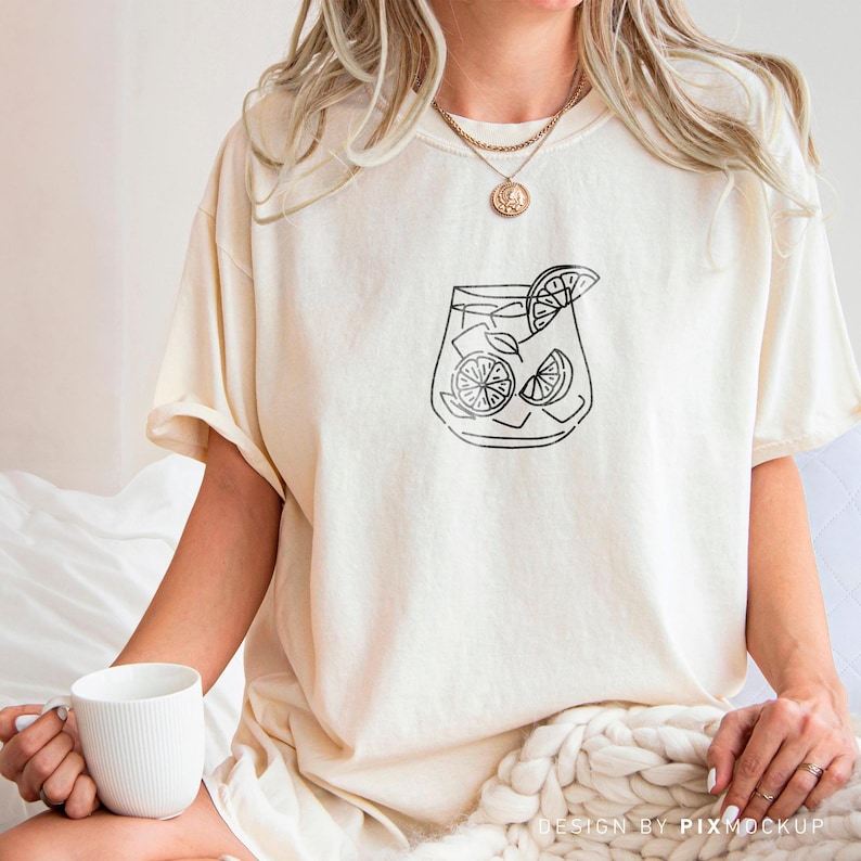 May include: A white t-shirt with a black line drawing of a glass of lemonade with lemon slices. The shirt is being worn by a person.