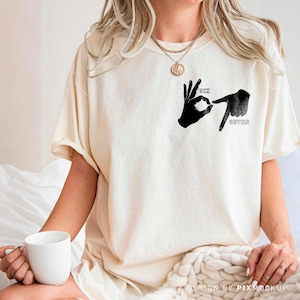 May include: Cream-colored t-shirt featuring a black graphic of two hands forming the numbers six and seven in sign language. The words "SIX" and "SEVEN" are printed above and below the hands. The shirt has a relaxed fit.