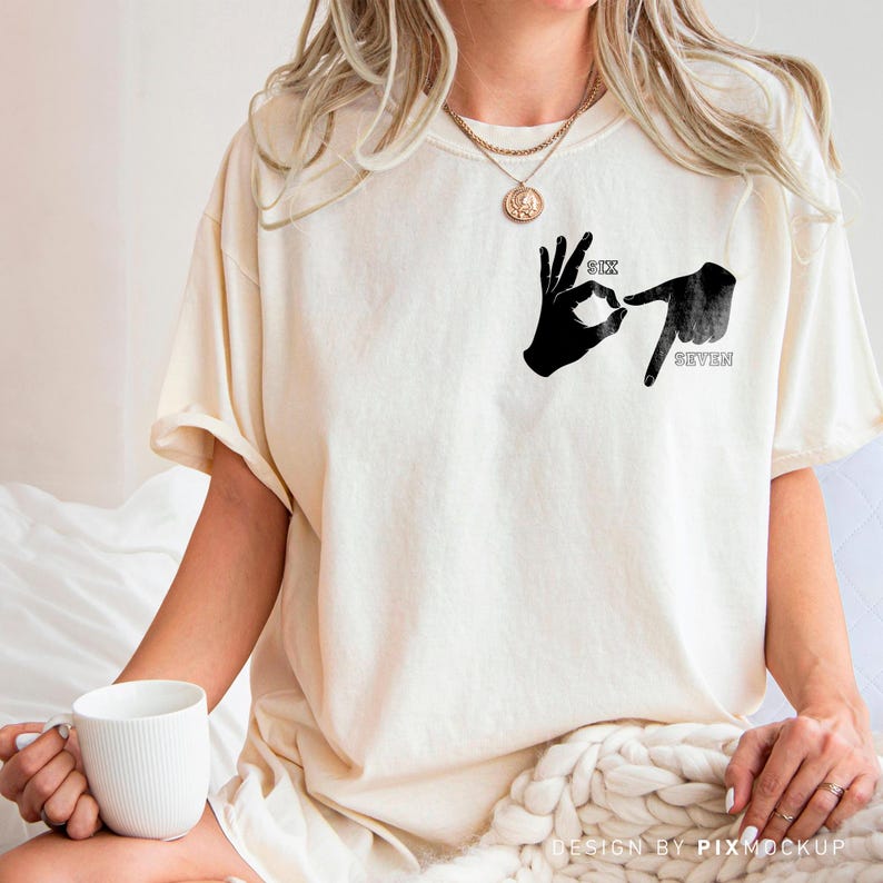 May include: Cream-colored t-shirt featuring a black graphic of two hands forming the numbers six and seven in sign language. The words "SIX" and "SEVEN" are printed above and below the hands. The shirt has a relaxed fit.