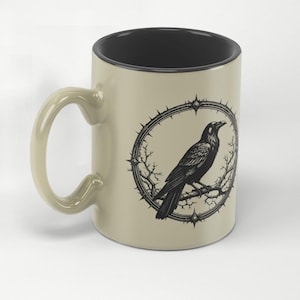 May include: A beige ceramic mug with a black interior and handle. The mug features a black illustration of a raven perched on a branch within a thorny circular frame. The mug has a light beige exterior.