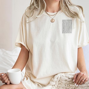 May include: Cream-colored t-shirt with a black vertical line design on the upper left chest. The shirt has short sleeves and a relaxed fit. A white mug is held in the hand.