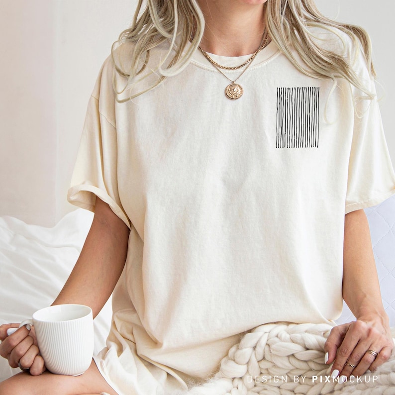 May include: Cream-colored t-shirt with a black vertical line design on the upper left chest. The shirt has short sleeves and a relaxed fit. A white mug is held in the hand.