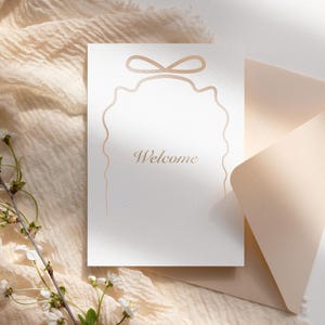 May include: A white card with a decorative border and the word "Welcome" in gold lettering. The card is on top of a light brown envelope and a piece of fabric.