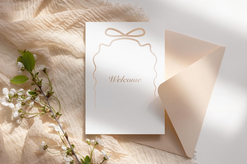 May include: A white card with a decorative border and the word "Welcome" in gold lettering. The card is on top of a light brown envelope and a piece of fabric.