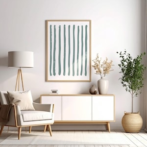 May include: A framed abstract art print featuring vertical teal brushstrokes on a white background. The artwork hangs above a white cabinet with wooden legs, complemented by a neutral-toned armchair, a floor lamp, and decorative plants, creating a modern interior design.