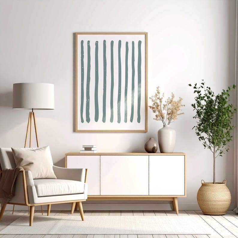 May include: A framed abstract art print featuring vertical teal brushstrokes on a white background. The artwork hangs above a white cabinet with wooden legs, complemented by a neutral-toned armchair, a floor lamp, and decorative plants, creating a modern interior design.