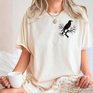 May include: Cream-colored t-shirt featuring a black raven perched on a tree branch. The graphic is on the left chest area. The shirt has short sleeves and a relaxed fit. The model is wearing a gold necklace and holding a white mug.
