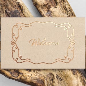 May include: A brown card with a gold foil design of a decorative frame and the word "Welcome" in the center.