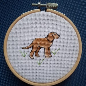May include: A brown dog cross-stitched on white fabric in a wooden embroidery hoop. The dog is standing on green grass.