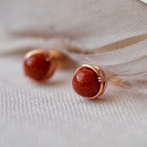 May include: A pair of small, round, brown gemstone earrings with a gold wire setting. The earrings are on a white fabric background.