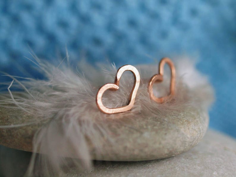 Puede incluir: Dos delicados pendientes en forma de coraz&oacute;n de oro rosa sobre una pluma blanca y una piedra gris.