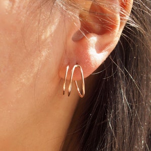 May include: A pair of gold hoop earrings with a unique, overlapping design. The earrings are worn in the left ear of a person.