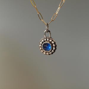 Peut inclure: Un collier pendentif rond avec une pierre précieuse bleue. Le pendentif argenté est orné d'un bord perlé et est suspendu à une chaîne dorée. Le bijou est mis en valeur sur un fond neutre.