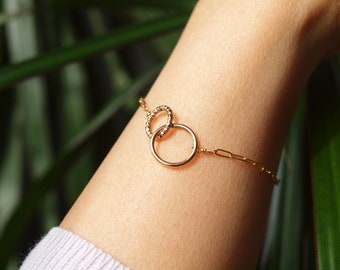 May include: A gold chain bracelet with two interlocking circles. The larger circle is plain, while the smaller circle has a textured surface.