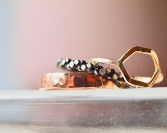 May include: A collection of rings in different styles. The base ring is copper-toned with a hammered finish. Above it is a ring with small black beads, and a gold ring with a geometric hexagon shape.
