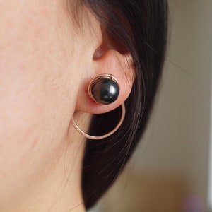 May include: A pair of earrings with a black pearl and a gold hoop. The earrings are worn in the ear of a person with dark hair.