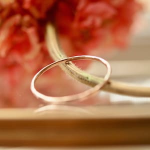 Peut inclure: Une fine bague en or, posée sur une surface en bois avec un arrière-plan flou de fleurs roses.