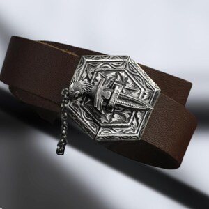 May include: A brown leather bracelet with a silver, ornate, hexagonal charm featuring a stylized dagger and a chain. The charm has intricate details and a textured surface.