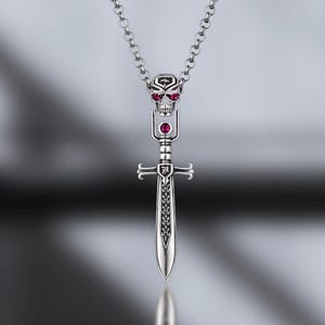 Könnte beinhalten: Silberne Anhänger-Halskette mit einem Schwertdesign und einem Totenkopf an der Spitze, verziert mit roten Edelsteinen. Der Schwertanhänger hat detaillierte Muster und hängt an einer feinen Kette. Die Halskette wird auf einer reflektierenden Oberfläche gezeigt.