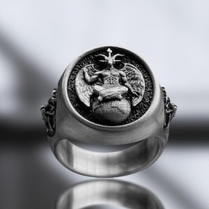 May include: A silver ring with a detailed depiction of a winged figure sitting on a sphere. The ring has a textured band and a raised, circular design.