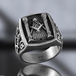 May include: A silver Masonic ring with a detailed design featuring two pillars, a compass and square, and a checkered floor. The ring is engraved with the number "925" on the inside band.