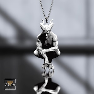 May include: A silver-toned pendant necklace featuring a detailed sculpture of a horned humanoid figure. The figure is crouched, with arms crossed, and rests on a clawed base. The pendant hangs from a delicate chain. The image is in black and white.