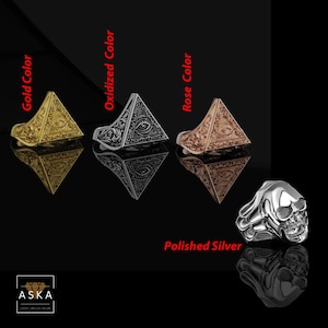 Op de afbeelding: Een collectie ringen in goud, geoxideerd en roze, plus een gepolijste zilveren schedelring. De ringen hebben piramide-ontwerpen met een oogsymbool. De ringen worden gepresenteerd op een reflecterend zwart oppervlak.