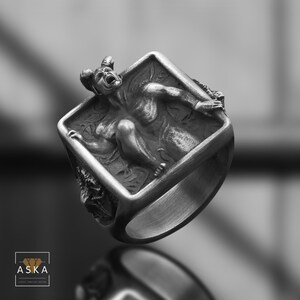 Può includere: Un anello d'argento con un designo dettagliato di un demone intrappolato in una cornice quadrata. Il demone è raffigurato con corna, ali e un'espressione di angoscia.